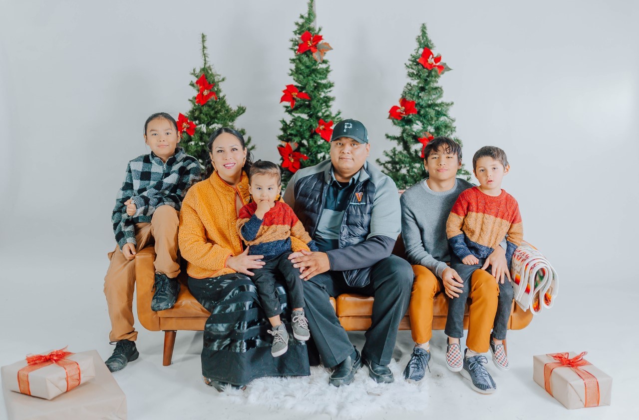 A family sitting on a couch in front of three Christmas trees.